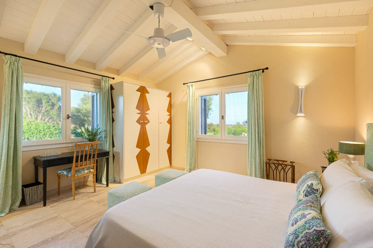 Bright and airy bedroom featuring a king-size bed with decorative pillows, a stylish black writing desk with a wooden chair, and a modern wardrobe with a geometric design. Large windows with green curtains provide natural light and a view of the outdoors, complemented by a ceiling fan and elegant wall lighting. | Only on SopranoVillas