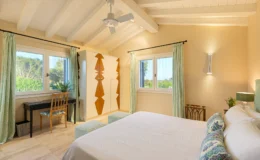 Bright and airy bedroom featuring a king-size bed with decorative pillows, a stylish black writing desk with a wooden chair, and a modern wardrobe with a geometric design. Large windows with green curtains provide natural light and a view of the outdoors, complemented by a ceiling fan and elegant wall lighting.