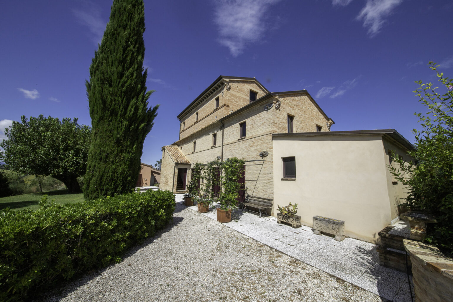 Historic stone villa surrounded by lush greenery and cypress trees, featuring a gravel pathway and a clear blue sky. Ideal for vacation rentals or countryside retreats. | Only on SopranoVillas
