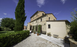 Historic stone villa surrounded by lush greenery and cypress trees, featuring a gravel pathway and a clear blue sky. Ideal for vacation rentals or countryside retreats.
