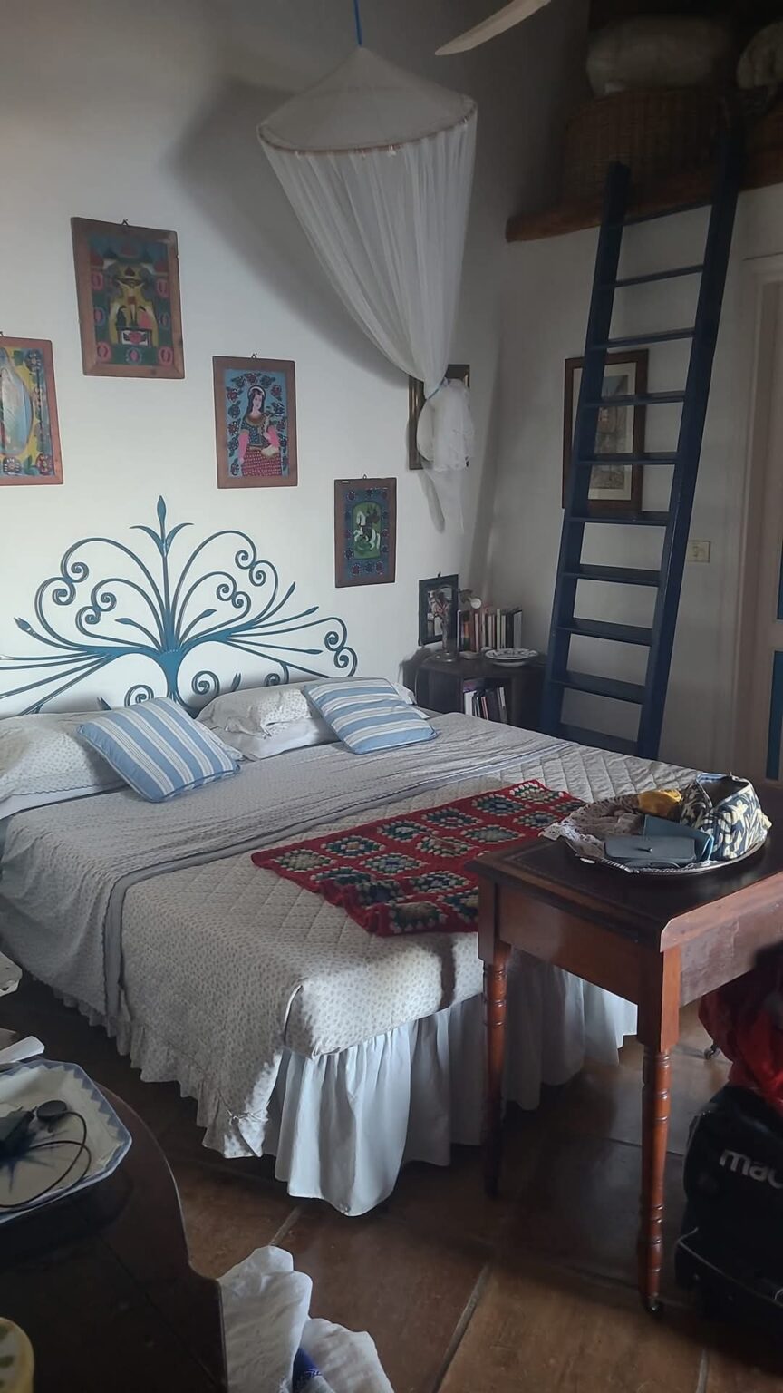 Cozy bedroom featuring a double bed with a decorative headboard, blue and white striped pillows, and a colorful blanket. The room is adorned with vibrant artwork on the walls and has a wooden table with various items. A blue ladder leads to a loft area, and a mosquito net hangs above the bed, adding charm to the space. | Only on SopranoVillas