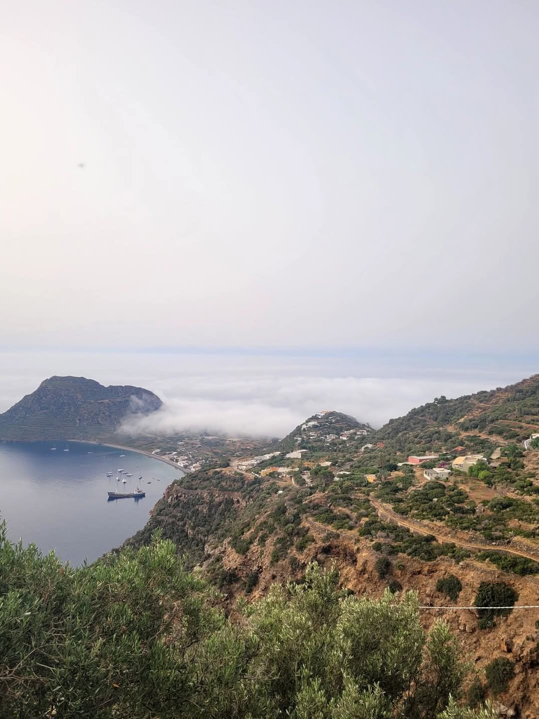 A panoramic view of a coastal landscape featuring a serene bay, dotted with boats, and surrounded by lush green hills. Low-lying clouds drift over the water, creating a misty atmosphere. The image captures the natural beauty and tranquility of the area, highlighting the blend of land and sea. | Only on SopranoVillas