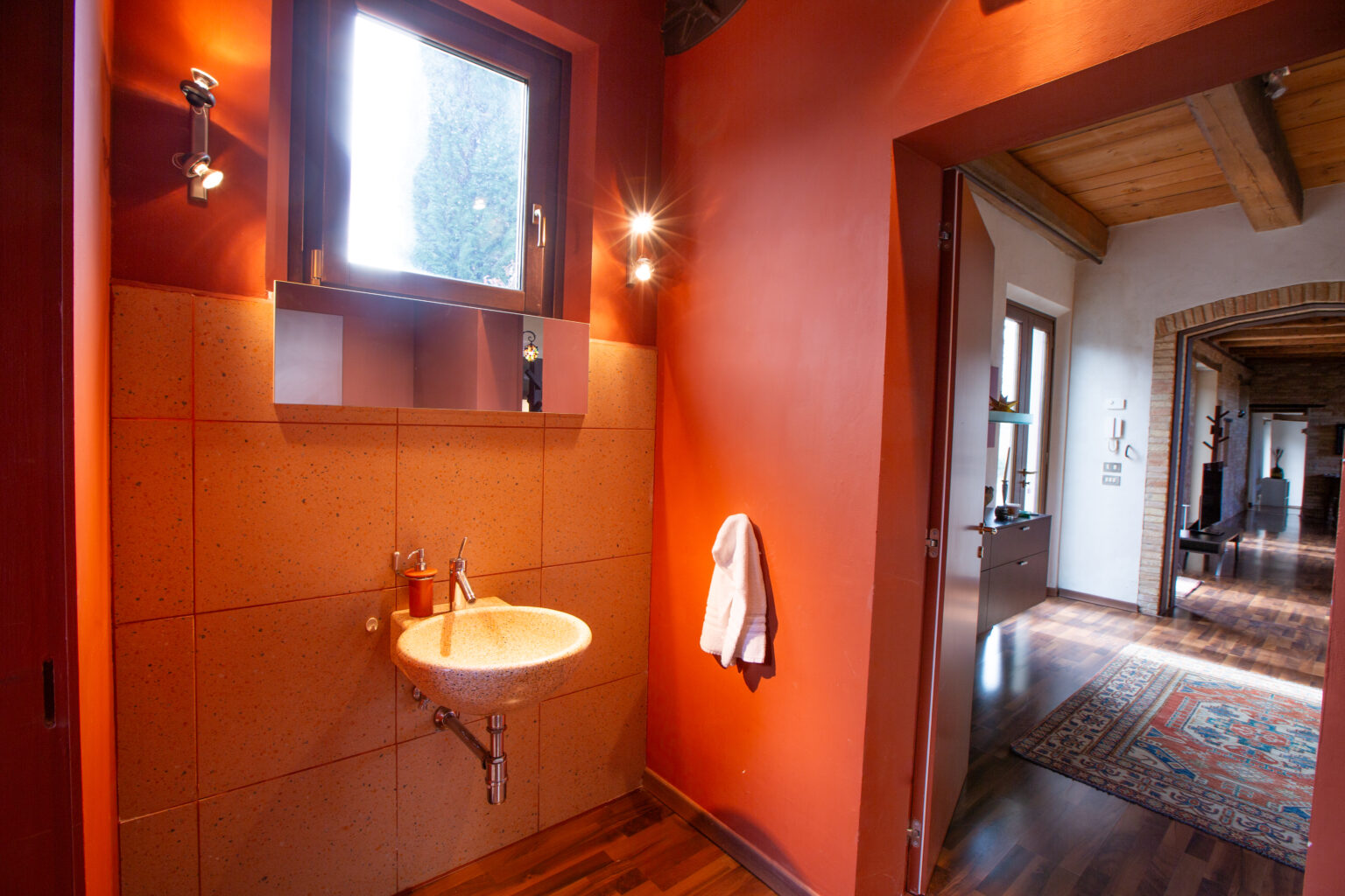 Modern bathroom with orange walls, a small sink, and a window, featuring warm lighting and a cozy atmosphere, leading to a hallway with wooden flooring and decorative elements. | Only on SopranoVillas
