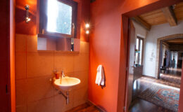 Modern bathroom with orange walls, a small sink, and a window, featuring warm lighting and a cozy atmosphere, leading to a hallway with wooden flooring and decorative elements.