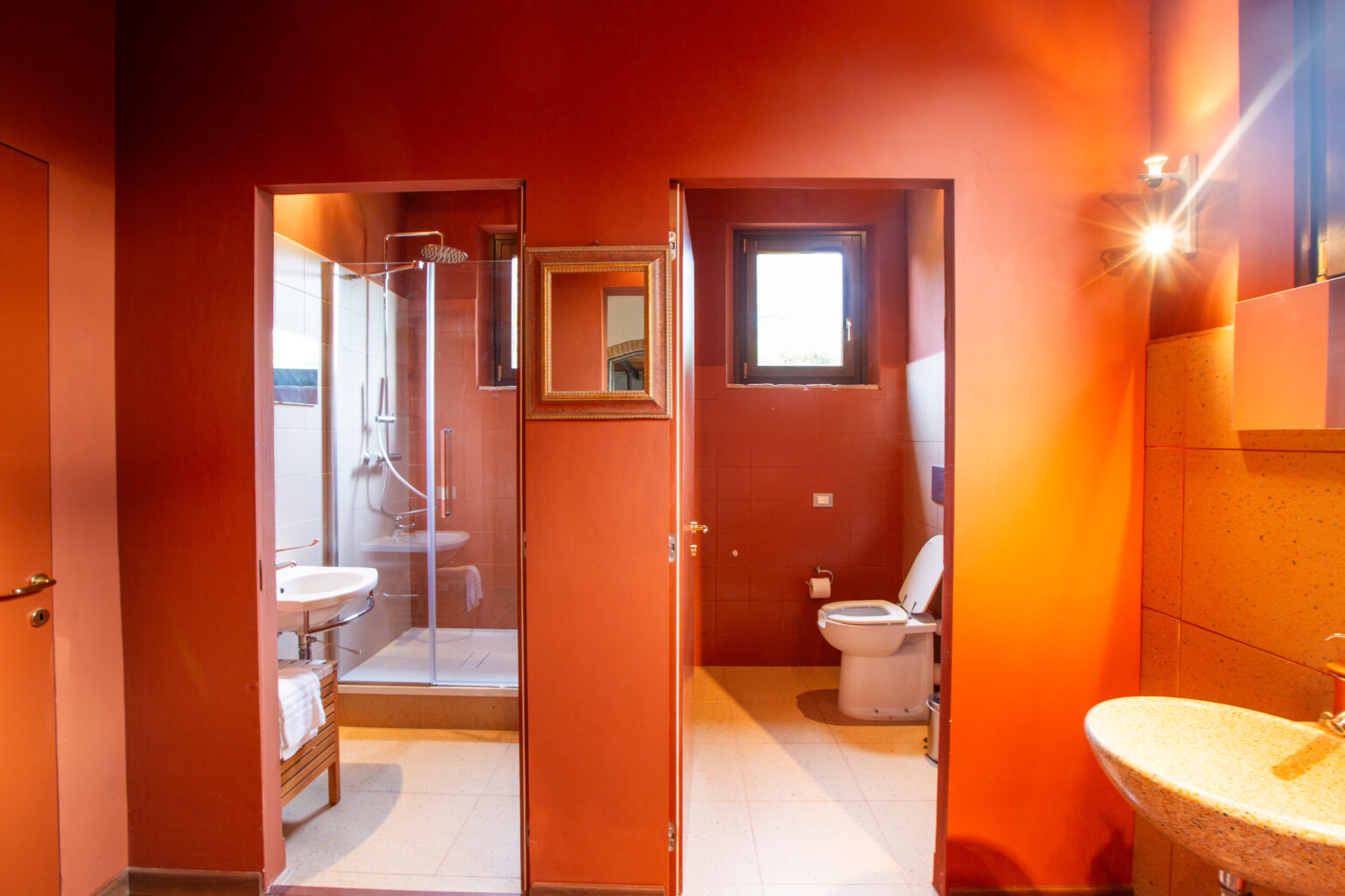 Modern bathroom interior featuring vibrant orange walls, a glass shower enclosure, a toilet, and a stylish sink. The layout emphasizes space and contemporary design elements, ideal for home improvement inspiration. | Only on SopranoVillas