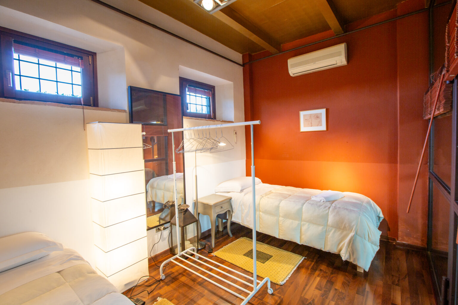 Cozy bedroom featuring two single beds, warm wooden flooring, and a stylish red accent wall. The room includes natural light from windows, a modern lamp, a clothing rack, and a mirror, creating a welcoming atmosphere for relaxation or work. | Only on SopranoVillas