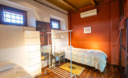 Cozy bedroom featuring two single beds, warm wooden flooring, and a stylish red accent wall. The room includes natural light from windows, a modern lamp, a clothing rack, and a mirror, creating a welcoming atmosphere for relaxation or work.