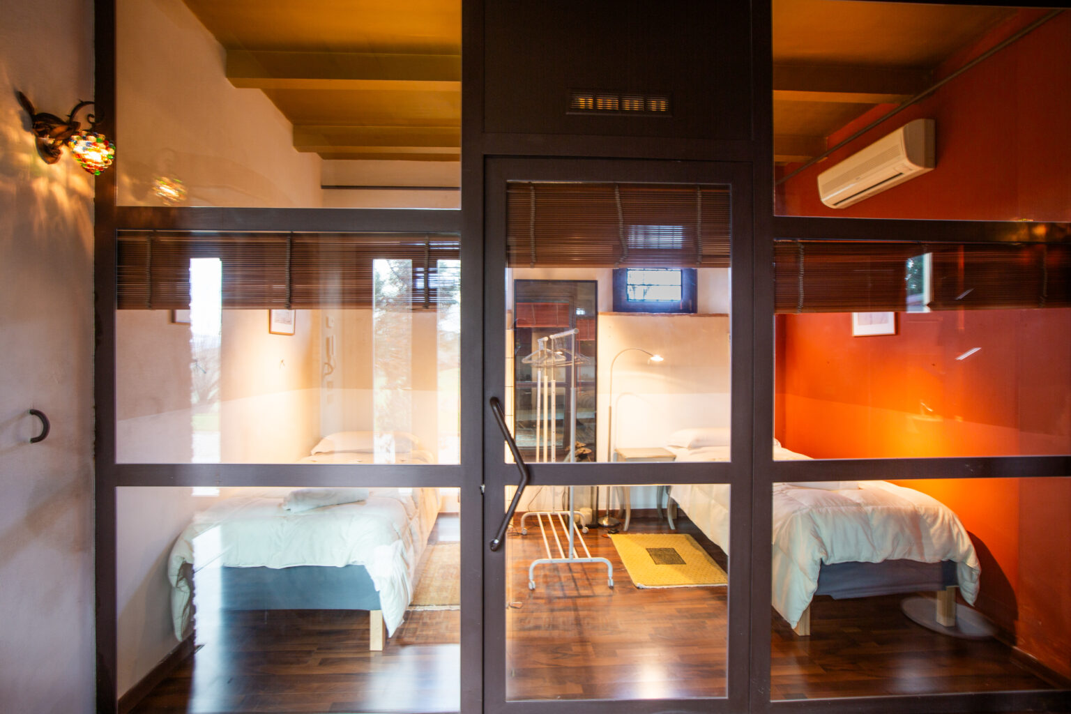 Modern shared accommodation featuring two twin beds, wooden flooring, and a glass partition. The room is well-lit with warm colors and includes air conditioning, creating a comfortable living space. Ideal for travelers seeking budget-friendly lodging. | Only on SopranoVillas
