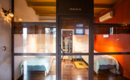 Modern shared accommodation featuring two twin beds, wooden flooring, and a glass partition. The room is well-lit with warm colors and includes air conditioning, creating a comfortable living space. Ideal for travelers seeking budget-friendly lodging.