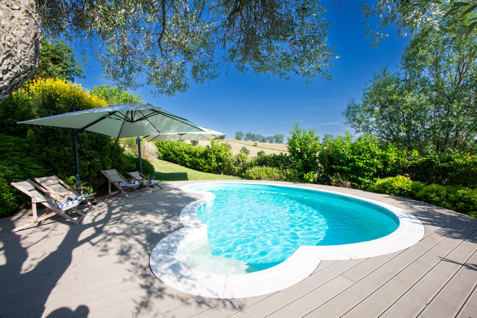 Serene outdoor pool area featuring a heart-shaped pool surrounded by lush greenery, wooden decking, and lounge chairs under a large umbrella, set against a clear blue sky. Ideal for relaxation and leisure. | Only on SopranoVillas