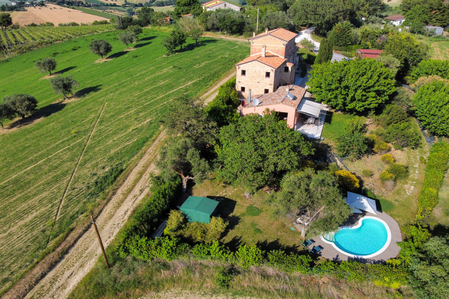 Aerial view of a charming countryside villa surrounded by lush greenery, featuring a swimming pool, landscaped garden, and nearby fields. Ideal for vacation rentals or serene retreats. | Only on SopranoVillas