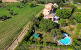 Aerial view of a charming countryside villa surrounded by lush greenery, featuring a swimming pool, landscaped garden, and nearby fields. Ideal for vacation rentals or serene retreats.