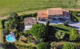 Aerial view of a spacious countryside villa with a pool, surrounded by lush greenery and a well-maintained lawn. The property features a rustic design and is situated in a serene landscape.