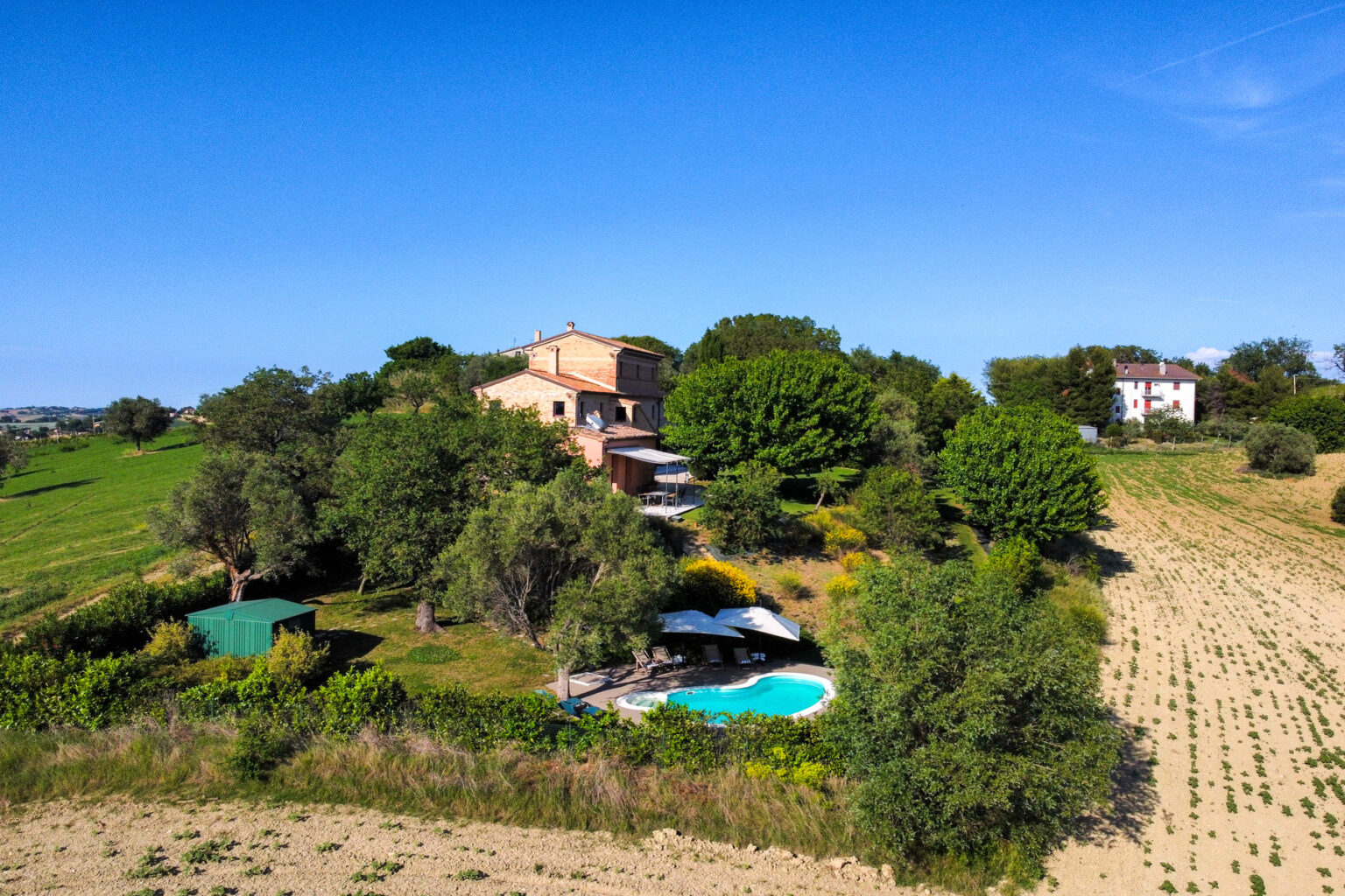 Aerial view of a picturesque countryside villa surrounded by lush greenery, featuring a swimming pool, shaded lounging areas, and open fields under a clear blue sky. Ideal for vacation rentals or serene getaways. | Only on SopranoVillas