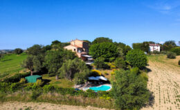 Aerial view of a picturesque countryside villa surrounded by lush greenery, featuring a swimming pool, shaded lounging areas, and open fields under a clear blue sky. Ideal for vacation rentals or serene getaways.