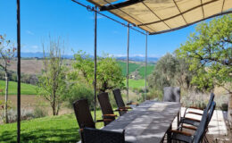 Outdoor dining area with a long table and chairs under a canopy, overlooking lush green fields and distant mountains on a clear day.