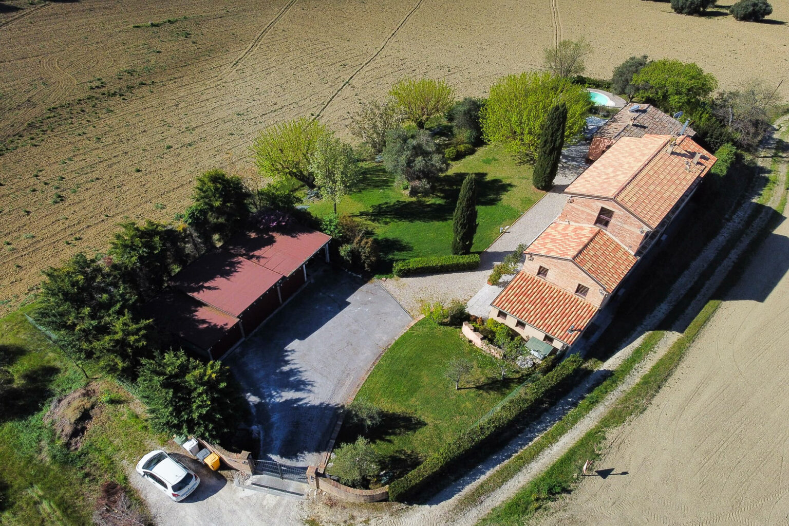 Aerial view of a picturesque countryside property featuring a spacious house with a red-tiled roof, surrounded by lush greenery and a well-maintained garden. A gravel driveway leads to a covered parking area, with open fields visible in the background. Ideal for rural living or a vacation retreat. | Only on SopranoVillas