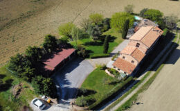 Aerial view of a picturesque countryside property featuring a spacious house with a red-tiled roof, surrounded by lush greenery and a well-maintained garden. A gravel driveway leads to a covered parking area, with open fields visible in the background. Ideal for rural living or a vacation retreat.