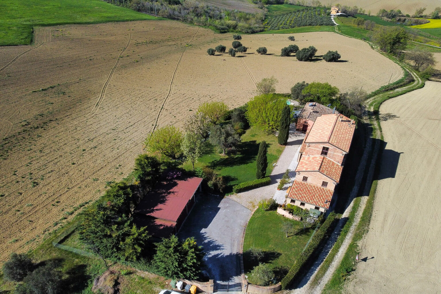 Aerial view of a rustic Italian farmhouse surrounded by lush greenery and farmland, featuring a red-roofed carport, well-maintained garden, and scenic rural landscape. | Only on SopranoVillas