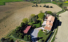 Aerial view of a rustic Italian farmhouse surrounded by lush greenery and farmland, featuring a red-roofed carport, well-maintained garden, and scenic rural landscape.