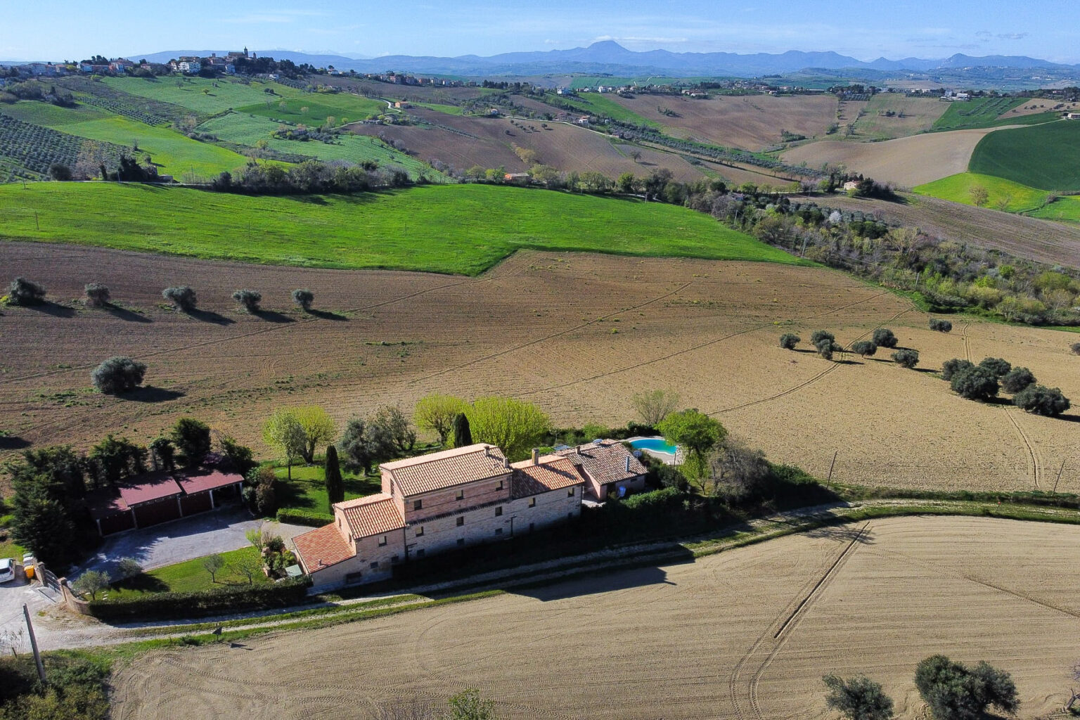 Aerial view of a charming countryside villa surrounded by lush green fields and rolling hills, showcasing a spacious outdoor area with a pool and landscaped gardens. Ideal for vacation rentals or country retreats. | Only on SopranoVillas