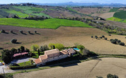 Aerial view of a charming countryside villa surrounded by lush green fields and rolling hills, showcasing a spacious outdoor area with a pool and landscaped gardens. Ideal for vacation rentals or country retreats.