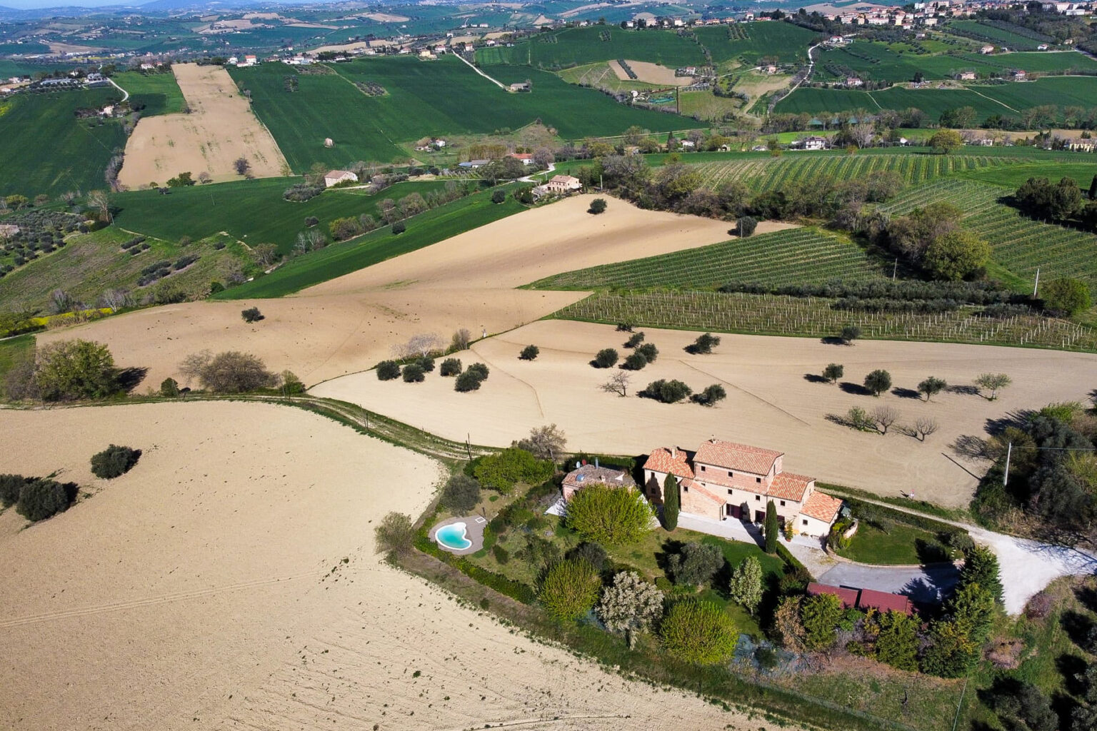 Aerial view of a rustic villa surrounded by rolling hills, lush green fields, and cultivated vineyards, with a small swimming pool and landscaped garden in the foreground. The scenic countryside landscape showcases a blend of agricultural land and natural beauty. | Only on SopranoVillas