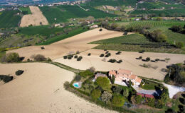 Aerial view of a rustic villa surrounded by rolling hills, lush green fields, and cultivated vineyards, with a small swimming pool and landscaped garden in the foreground. The scenic countryside landscape showcases a blend of agricultural land and natural beauty.