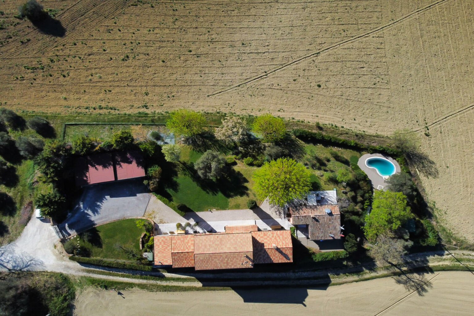Aerial view of a rural property featuring a spacious house with a red-tiled roof, surrounded by lush greenery and a swimming pool. The landscape includes farmland in the background, offering a serene and picturesque setting. | Only on SopranoVillas