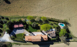 Aerial view of a rural property featuring a spacious house with a red-tiled roof, surrounded by lush greenery and a swimming pool. The landscape includes farmland in the background, offering a serene and picturesque setting.