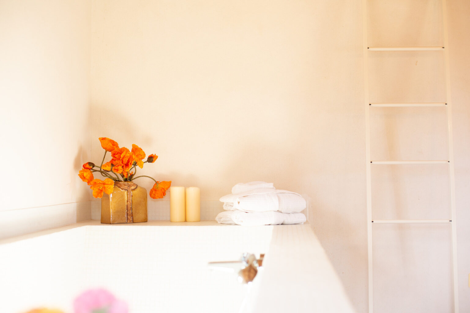 Bright and serene bathroom corner featuring a vase of orange flowers, two cream-colored candles, and neatly stacked white towels, with a minimalist ladder in the background. | Only on SopranoVillas