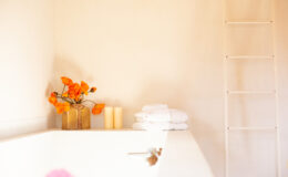 Bright and serene bathroom corner featuring a vase of orange flowers, two cream-colored candles, and neatly stacked white towels, with a minimalist ladder in the background.