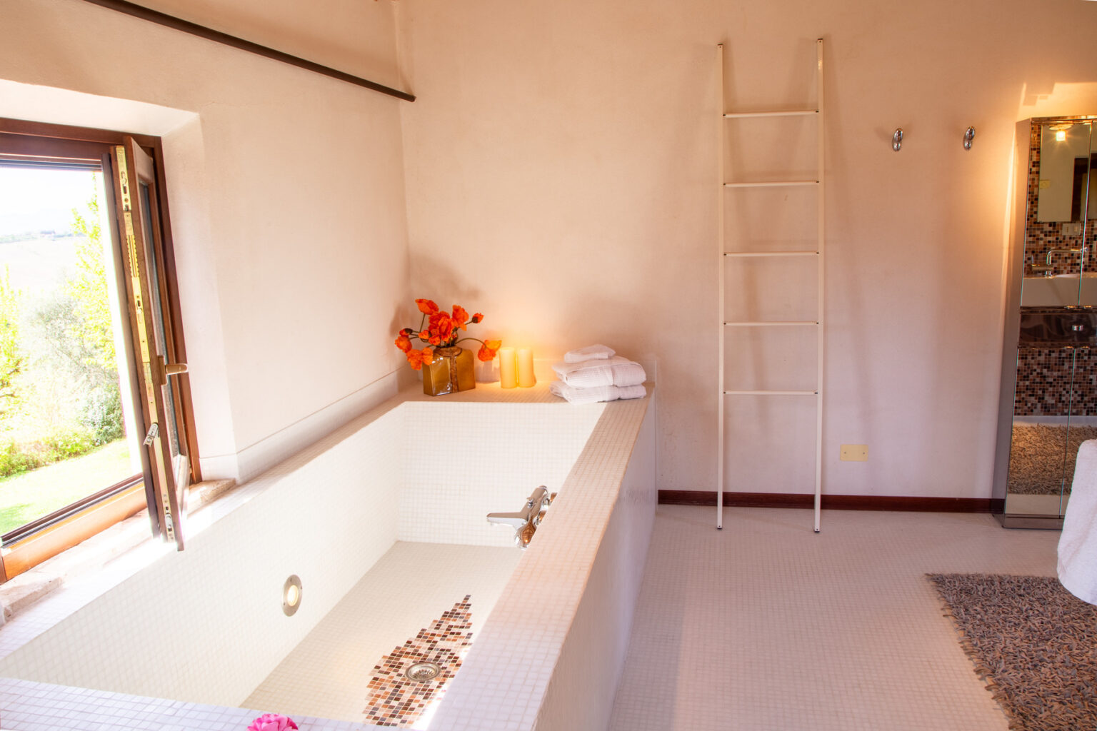 Modern bathroom interior featuring a spacious white tiled bathtub, orange floral arrangement, neatly stacked towels, and a decorative ladder. The room is illuminated by natural light from an open window, enhancing the serene atmosphere. | Only on SopranoVillas