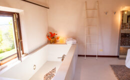 Modern bathroom interior featuring a spacious white tiled bathtub, orange floral arrangement, neatly stacked towels, and a decorative ladder. The room is illuminated by natural light from an open window, enhancing the serene atmosphere.