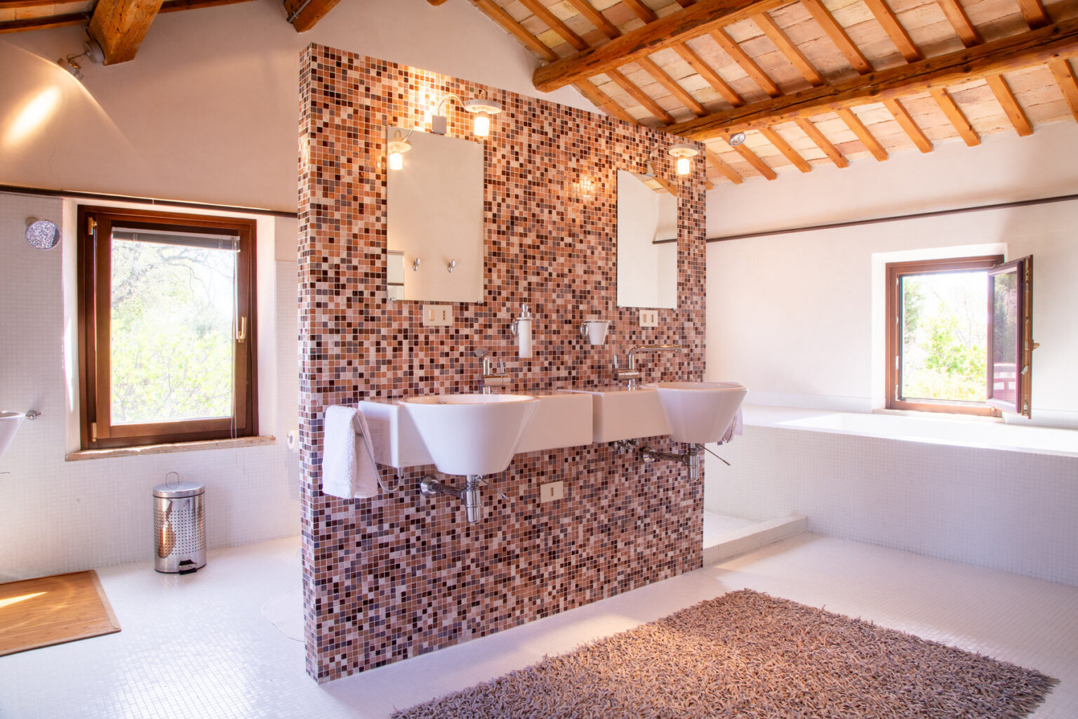 Modern bathroom featuring a unique cork wall design, double sinks, large mirrors, and natural light from windows, complemented by wooden beams and a cozy rug. | Only on SopranoVillas