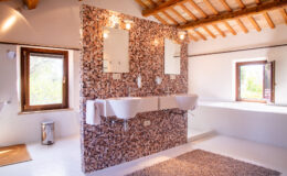 Modern bathroom featuring a unique cork wall design, double sinks, large mirrors, and natural light from windows, complemented by wooden beams and a cozy rug.
