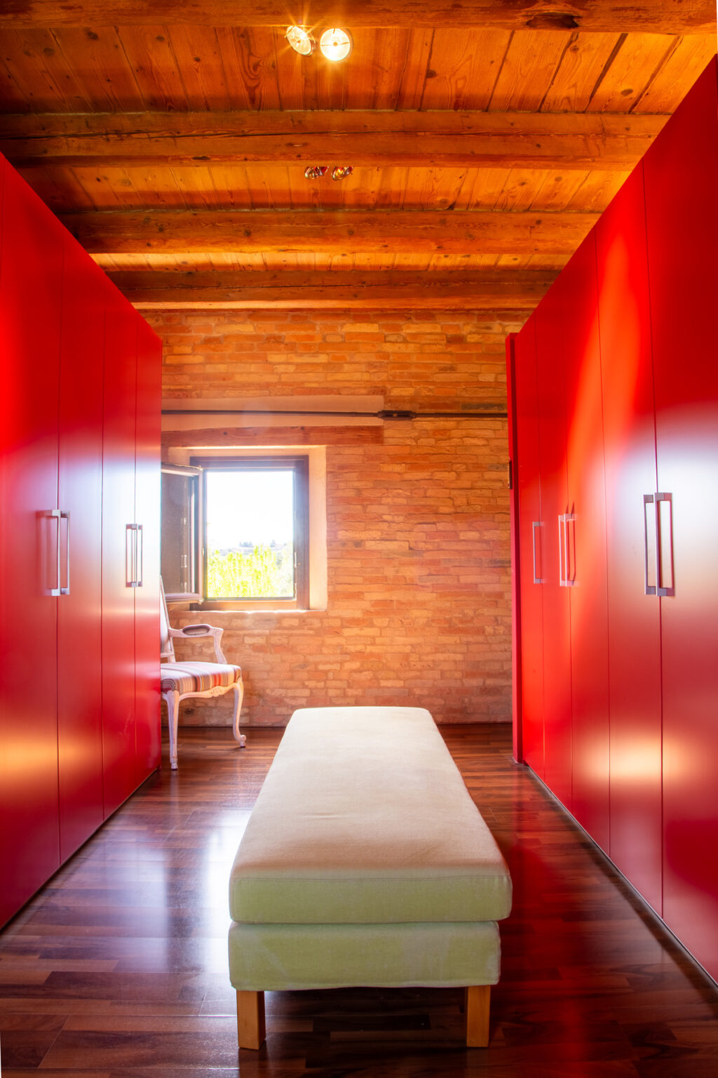 Modern interior design featuring a hallway with vibrant red wardrobes, a light-colored bench, and a window allowing natural light, set against a warm, wooden brick wall. | Only on SopranoVillas