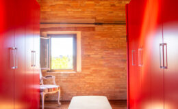 Modern interior design featuring a hallway with vibrant red wardrobes, a light-colored bench, and a window allowing natural light, set against a warm, wooden brick wall.