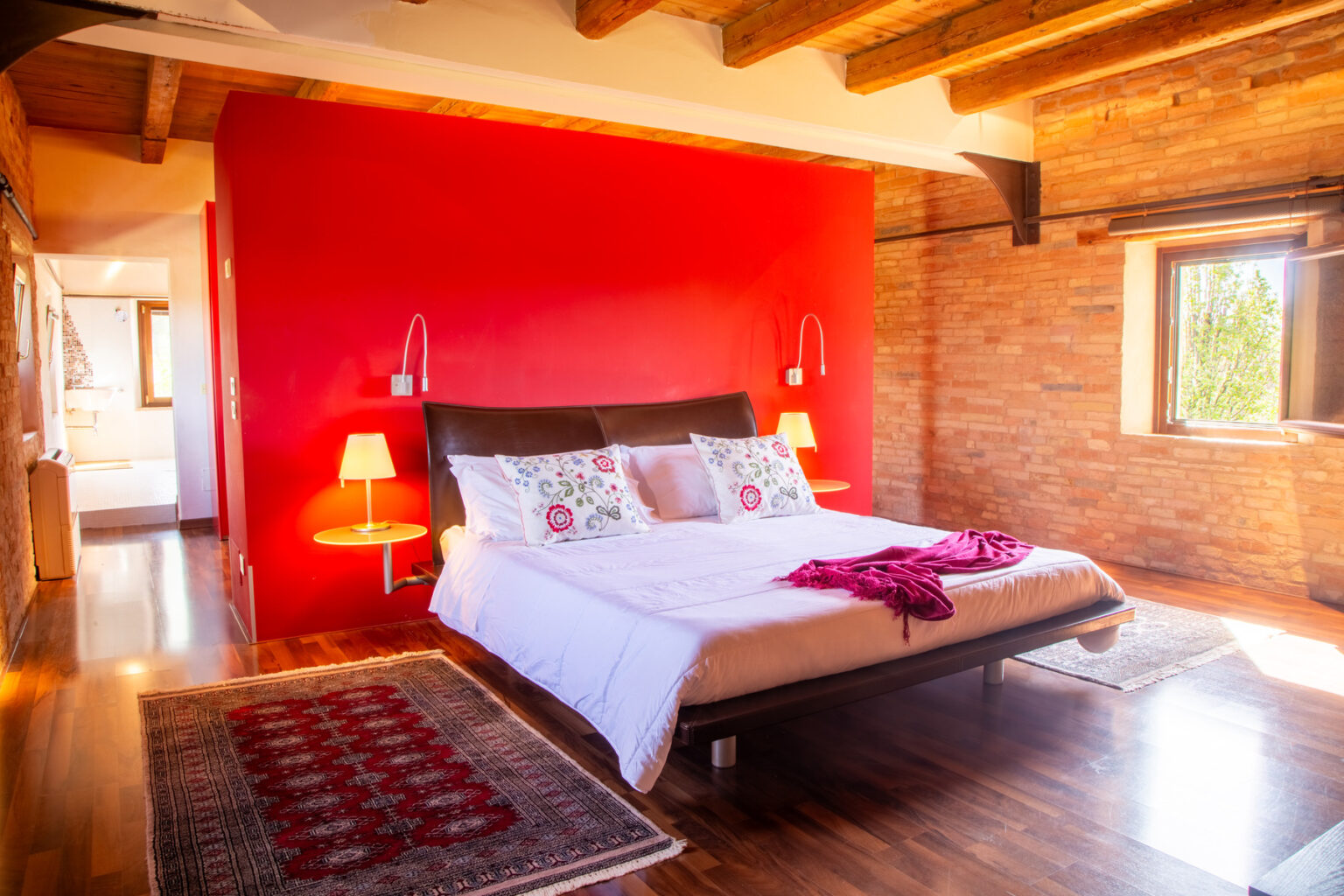 Cozy bedroom featuring a modern bed with decorative pillows, a vibrant red accent wall, wooden beams, and stylish lamps, set in a rustic environment with hardwood flooring. | Only on SopranoVillas