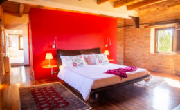 Cozy bedroom featuring a modern bed with decorative pillows, a vibrant red accent wall, wooden beams, and stylish lamps, set in a rustic environment with hardwood flooring.