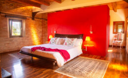 Cozy bedroom with a king-sized bed adorned with colorful pillows, a red accent wall, wooden beams, and warm lighting, featuring a stylish rug and decorative elements for a modern yet inviting atmosphere.