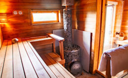 Interior of a modern sauna featuring wooden benches, a stone heater, and large windows for natural light, creating a relaxing atmosphere for wellness and rejuvenation.