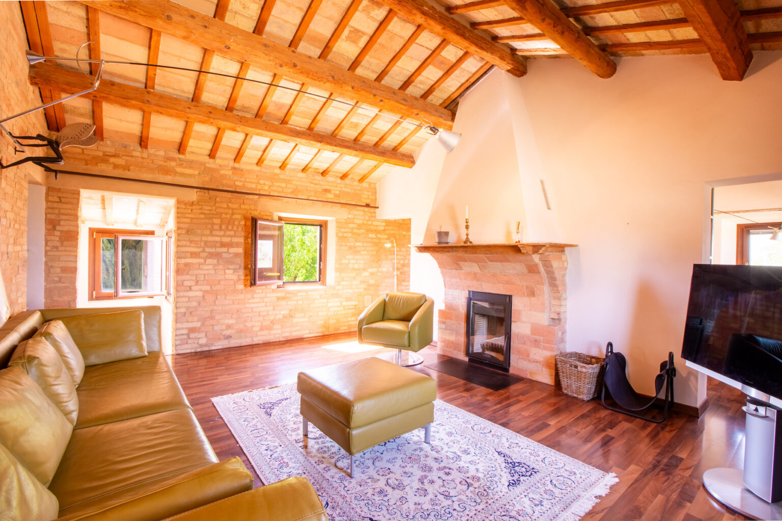 Cozy living room with wooden beams, stone walls, and a modern fireplace, featuring a green leather sofa, an ottoman, and a flat-screen TV, illuminated by natural light from windows. | Only on SopranoVillas