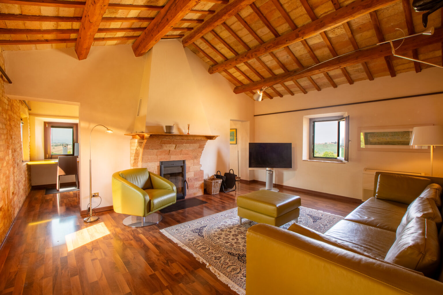 Cozy living room featuring wooden beams, a stone fireplace, and modern furnishings, including green armchairs and a flat-screen TV, with natural light streaming through windows and a view of the countryside. | Only on SopranoVillas
