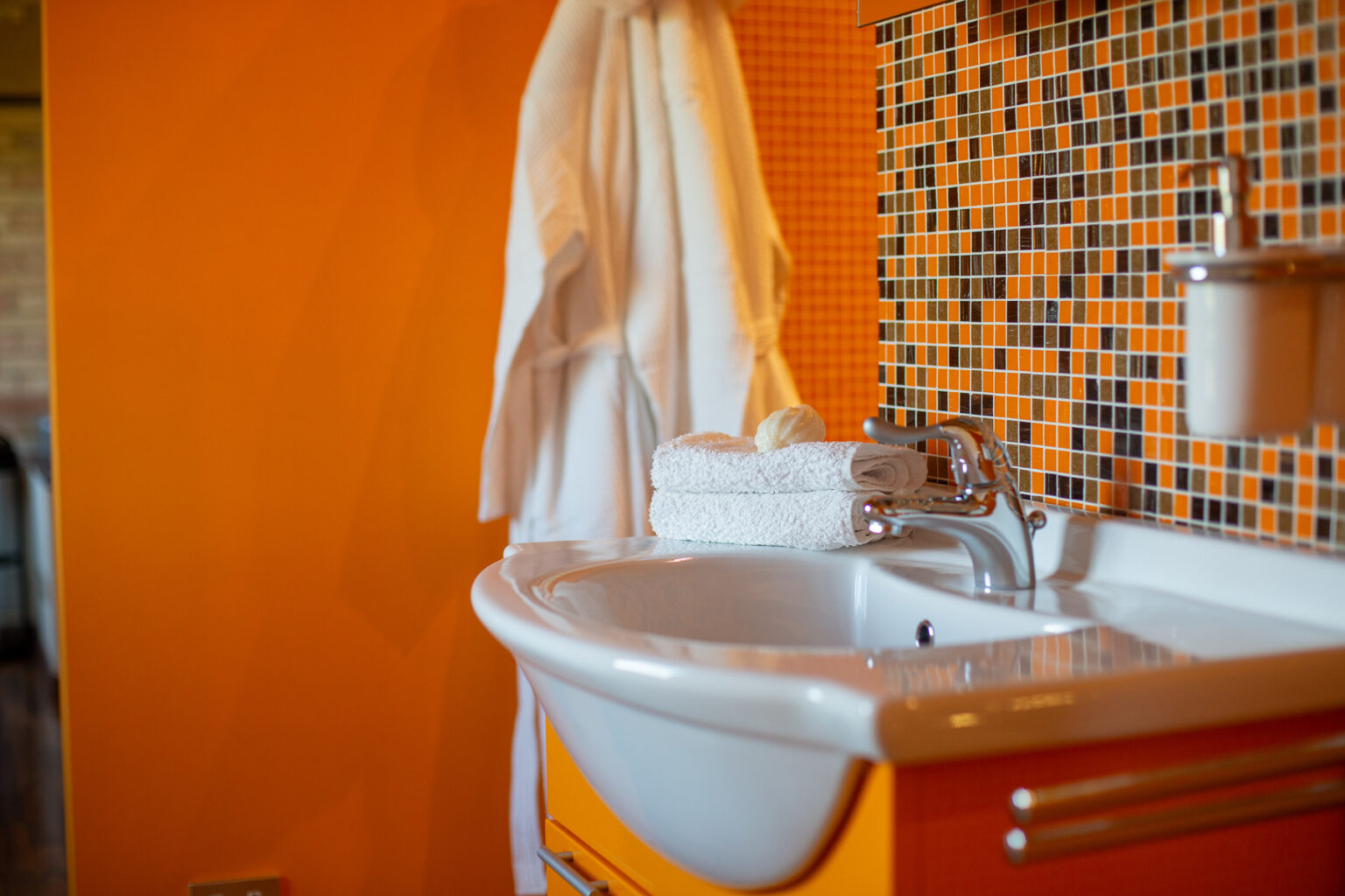 Modern bathroom with a vibrant orange wall, featuring a white sink, neatly stacked towels, and a bar of soap. A plush bathrobe hangs nearby, enhancing the inviting atmosphere. Ideal for showcasing contemporary interior design. | Only on SopranoVillas