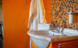 Modern bathroom with a vibrant orange wall, featuring a white sink, neatly stacked towels, and a bar of soap. A plush bathrobe hangs nearby, enhancing the inviting atmosphere. Ideal for showcasing contemporary interior design.