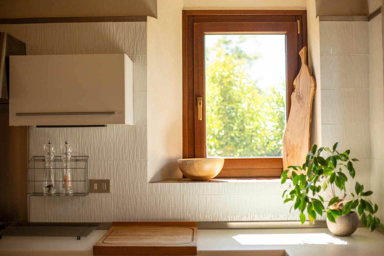 Bright and airy kitchen interior featuring a wooden window frame, a round wooden bowl on the windowsill, and a small potted plant. The wall displays textured tiles and a modern kitchen cabinet, with pepper and salt shakers neatly arranged in a wire holder. Natural light illuminates the space, enhancing its inviting atmosphere. | Only on SopranoVillas