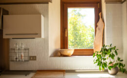 Bright and airy kitchen interior featuring a wooden window frame, a round wooden bowl on the windowsill, and a small potted plant. The wall displays textured tiles and a modern kitchen cabinet, with pepper and salt shakers neatly arranged in a wire holder. Natural light illuminates the space, enhancing its inviting atmosphere.