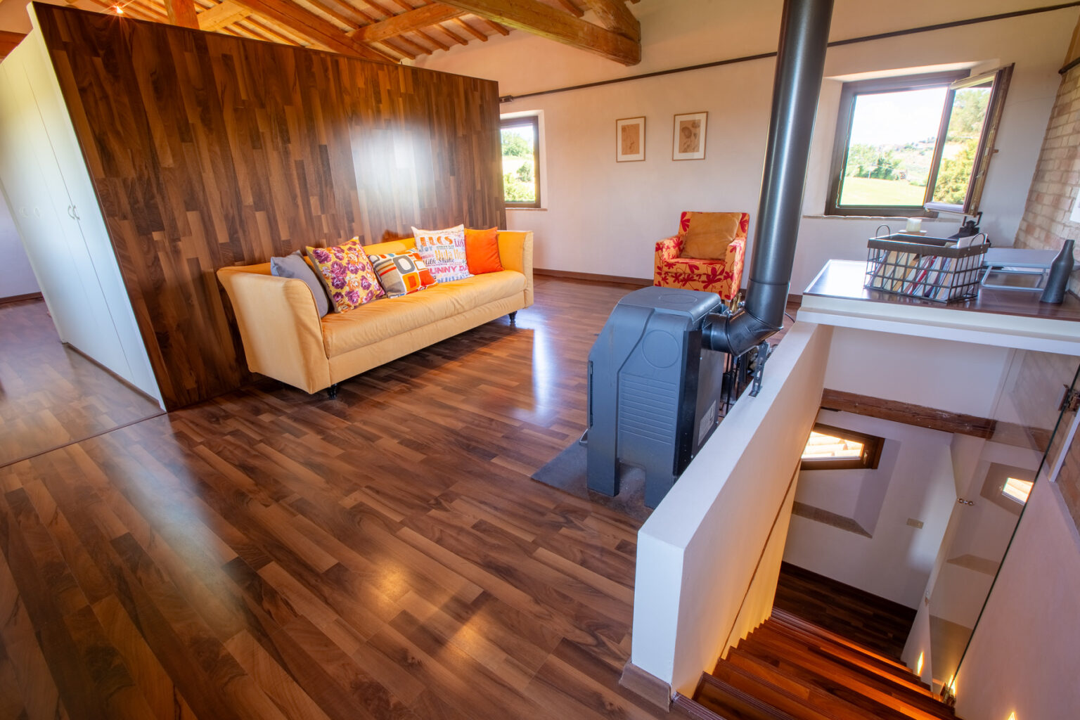 Cozy living room featuring a beige sofa with colorful cushions, a red patterned armchair, and a modern wood stove. The space has warm wooden flooring and large windows that offer a view of the countryside, creating a bright and inviting atmosphere. | Only on SopranoVillas