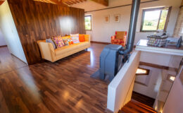 Cozy living room featuring a beige sofa with colorful cushions, a red patterned armchair, and a modern wood stove. The space has warm wooden flooring and large windows that offer a view of the countryside, creating a bright and inviting atmosphere.
